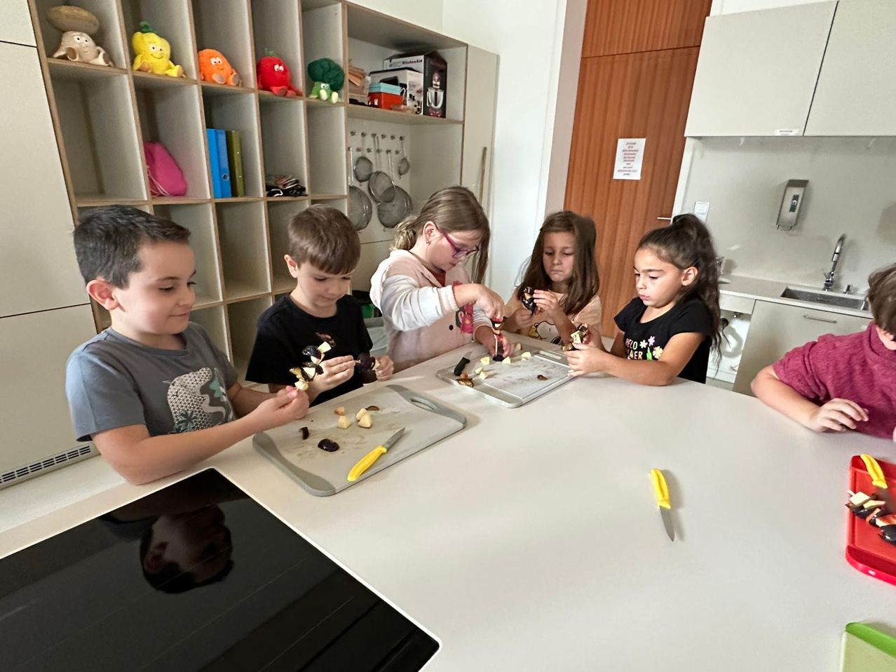 Fünf Kinder versammeln sich um einen Küchentisch und beschäftigen sich mit dem Schneiden von Gemüse. Jedes Kind hält ein Messer und schneidet verschiedene Gemüsesorten. Ein Schneidebrett und Messer liegen auf dem Tisch. Im Hintergrund sind Regale mit Spielzeug und Büchern zu sehen.