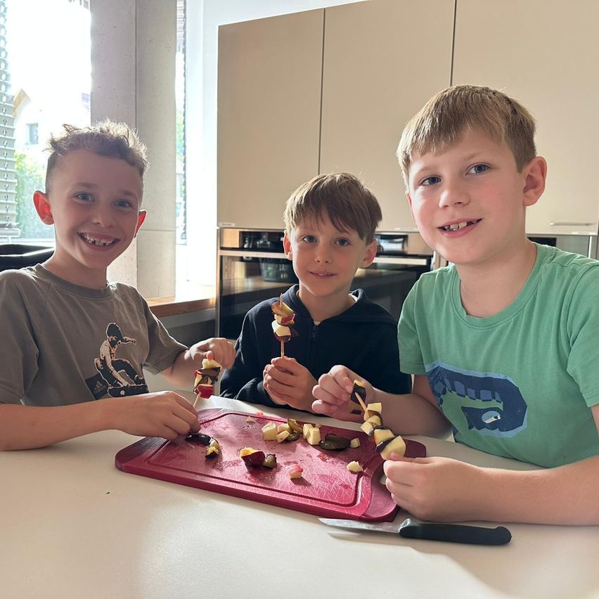 Drei Jungen sitzen lächelnd am Tisch und halten Obstspieße in der Hand. Der Junge auf der linken Seite trägt ein graues T-Shirt, der mittlere Junge einen schwarzen Pullover. Der Junge auf der rechten Seite trägt ein grünes T-Shirt. Sie sind von einer Küchenumgebung umgeben.