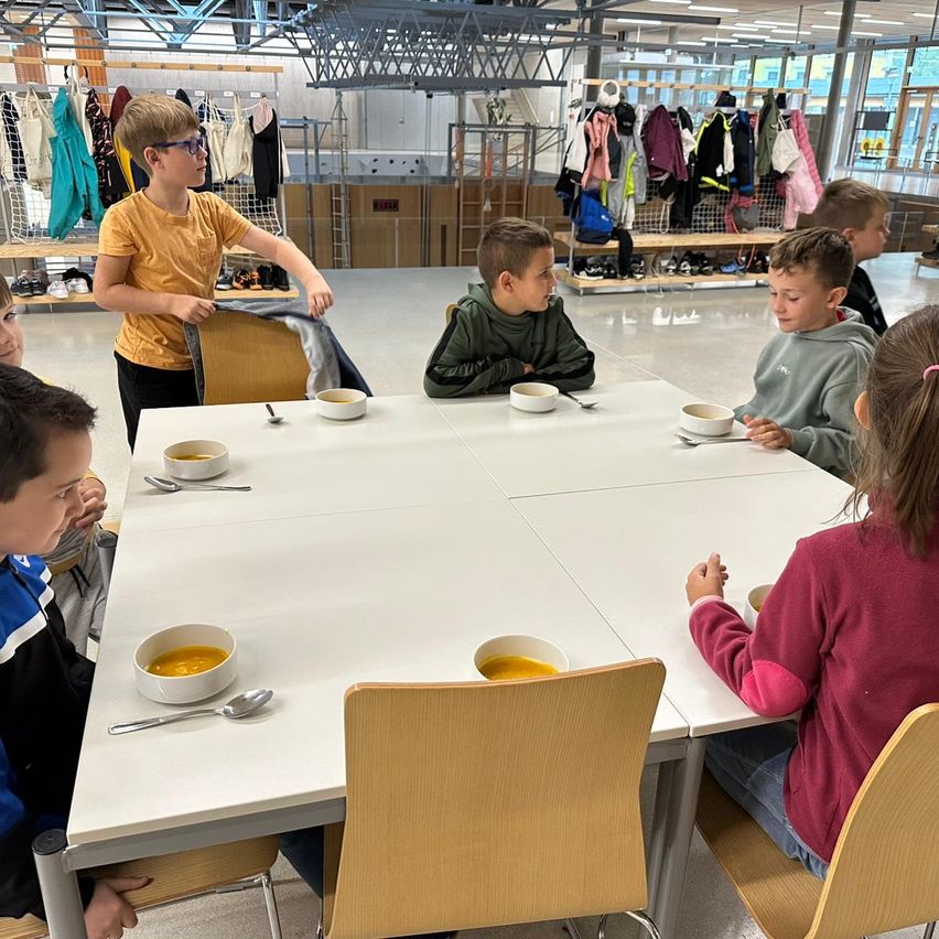 Eine Gruppe von Kindern sitzt um einen Tisch herum und isst Suppe, während ein Junge in der Nähe einen Mantel hält. Dahinter hängen Kleidungsstücke an Kleiderbügeln, und Schuhe sind auf Regalen ausgestellt.