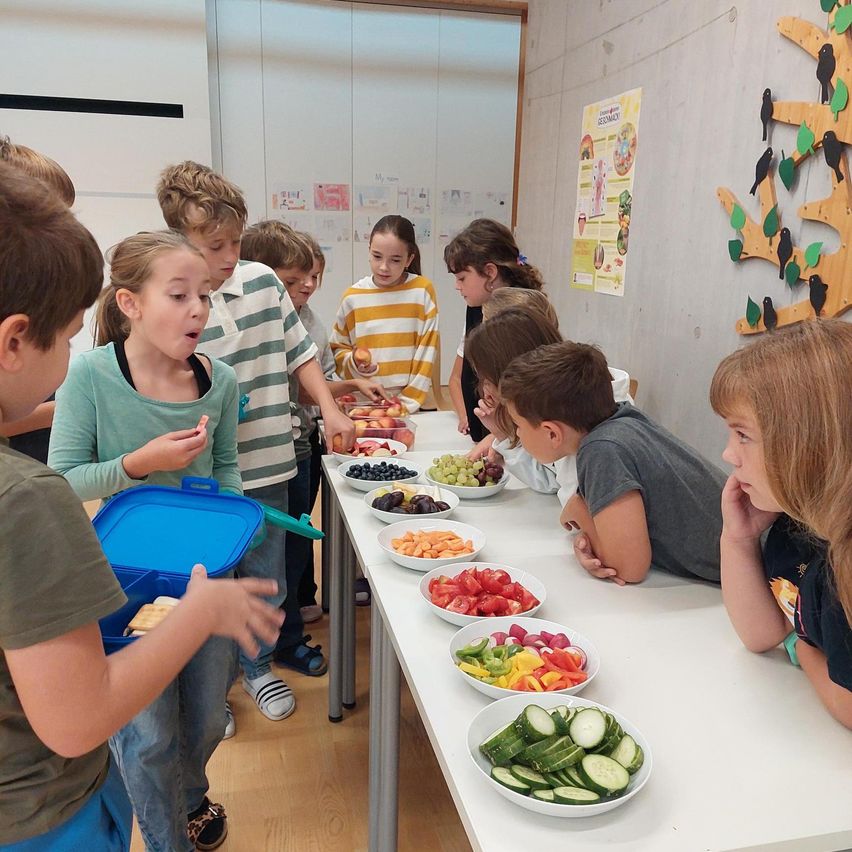 Eine Gruppe von Kindern steht um einen Tisch, der mit verschiedenen Obst- und Gemüsesorten gefüllt ist. Einige essen und andere schauen sich das Essen an.