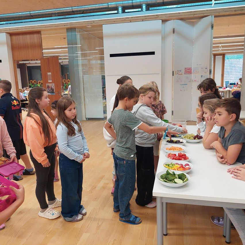 In einem Innenraum versammeln sich Kinder um einen Tisch voller verschiedenem Obst und Gemüse. Einige Kinder greifen nach dem Essen, während andere stehen und zusehen. Es gibt Glaswände und Türen und einen Holzboden.