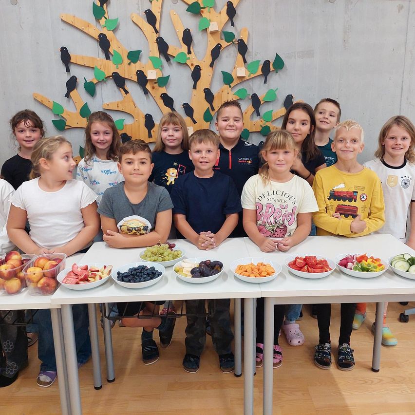 Eine Gruppe von Kindern steht hinter zwei Tischen mit Obst und Gemüse. Sie posieren für ein Foto in einem Raum mit einer Baumwand.