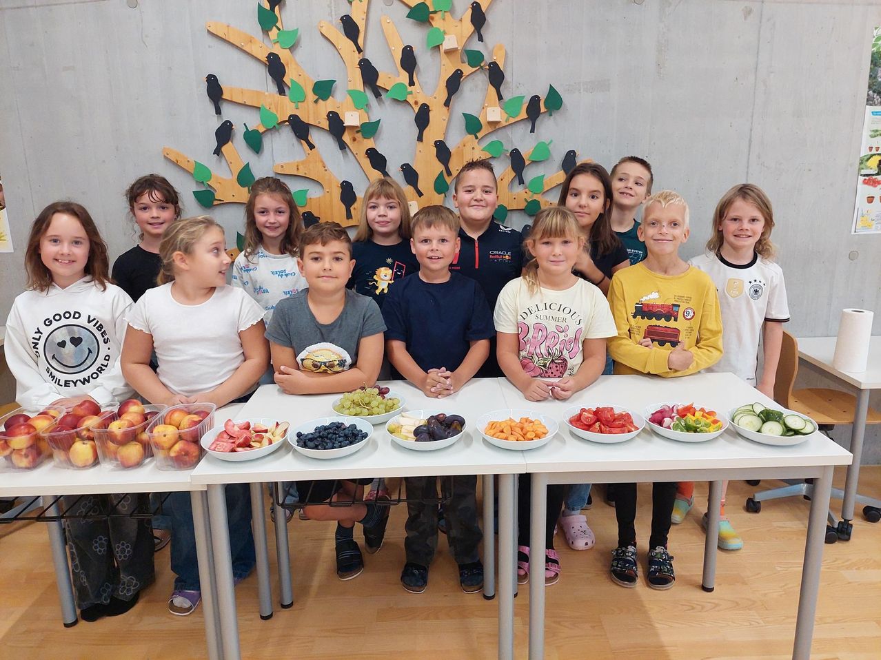 Eine Gruppe von Kindern steht hinter zwei Tischen mit Obst und Gemüse. Sie posieren für ein Foto in einem Raum mit einer Baumwand.