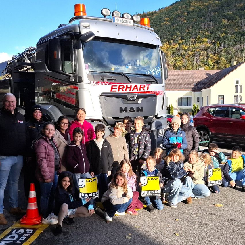 Eine Gruppe von Menschen posiert mit einem MAN-Lkw auf einer Straße und hält Schilder für Friends on the Road. Der Lkw hat das Wort ERHART auf der Vorderseite. Ein Verkehrskegel steht auf der Straße.