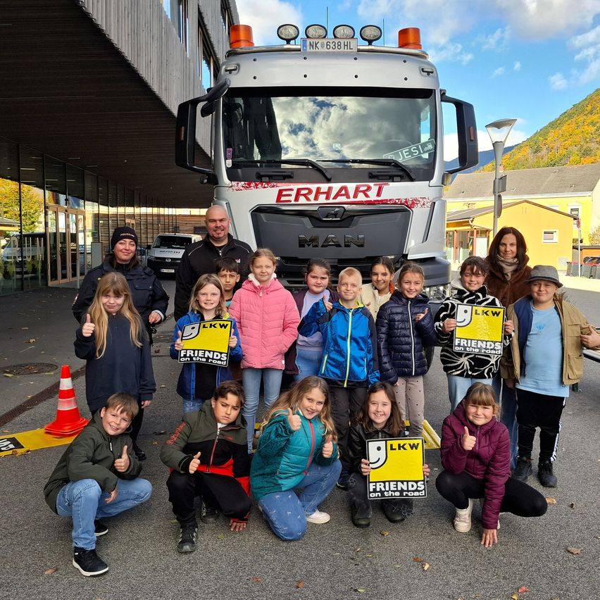 Gruppe von Kindern und Erwachsenen, die vor einem LKW posieren und Schilder mit der Aufschrift 'LKW Friends on the Road' halten. Dahinter steht ein Gebäude mit Glasfenstern und ein gelber LKW im Hintergrund.