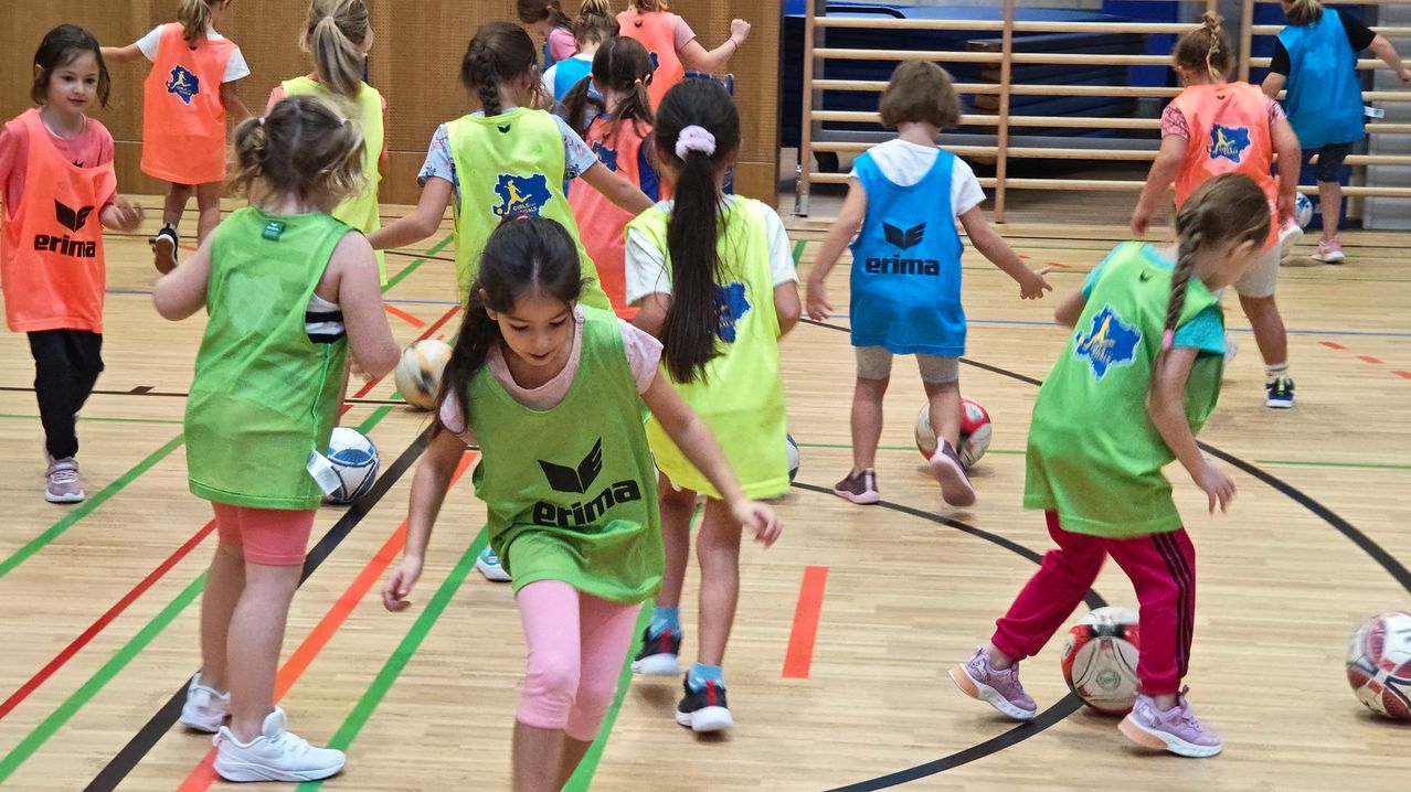 Eine Gruppe junger Mädchen in grünen Westen spielt Fußball auf einem Holzboden. Jedes Mädchen hält einen Fußball und rennt und jagt dem Ball hinterher. Im Hintergrund ist eine Holzleiter zu sehen.