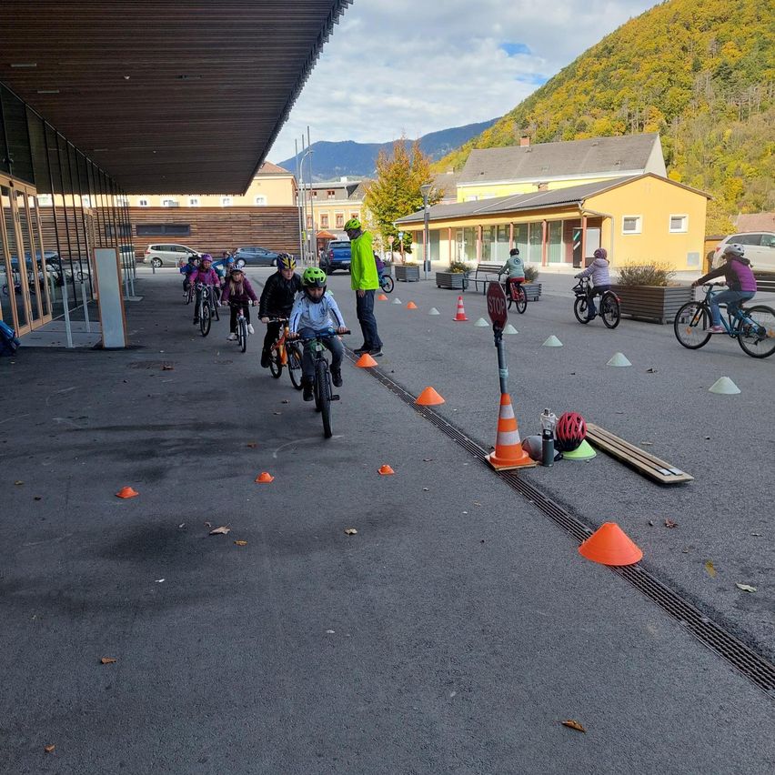 Kinder fahren in einer Reihe auf einem gepflasterten Bereich mit Verkehrskegeln und einem Stoppschild auf Fahrrädern. Gebäude und Berge sind im Hintergrund.