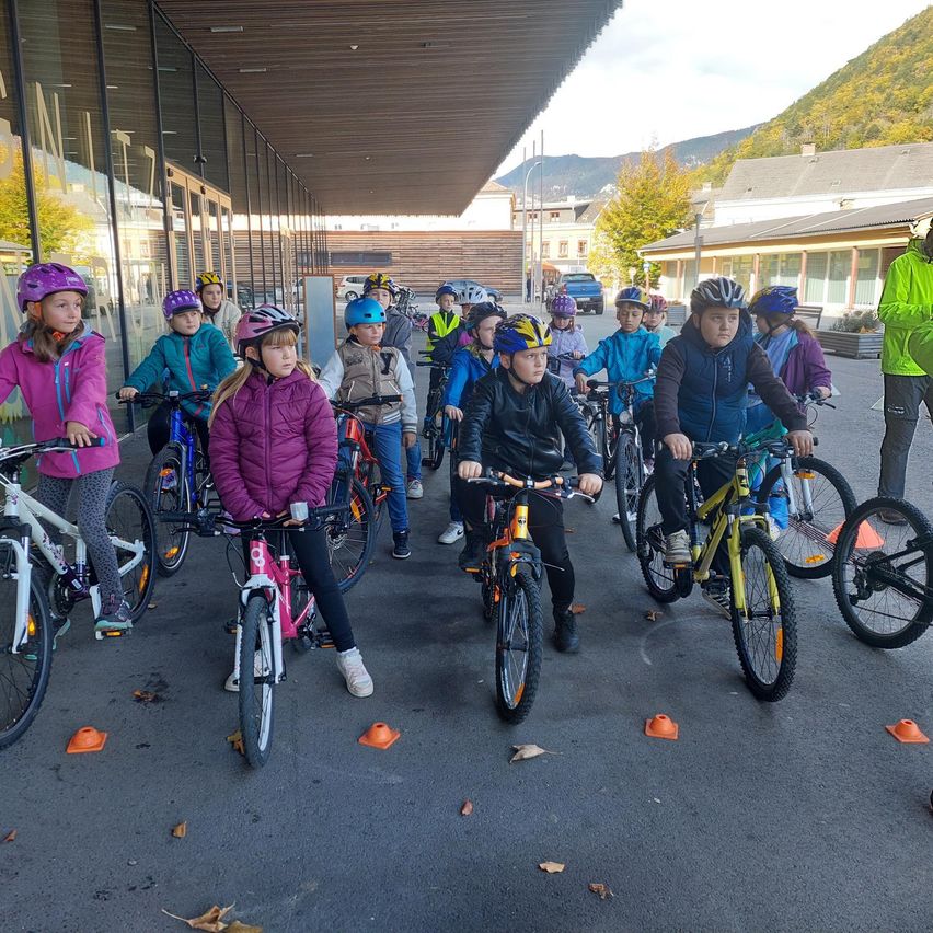 Eine Gruppe von Kindern ist auf Fahrrädern in einem Außenbereich versammelt und trägt Helme. Sie sind mit orangefarbenen Kegeln auf dem Boden aufgereiht. Dahinter steht ein Gebäude mit Glaswänden.