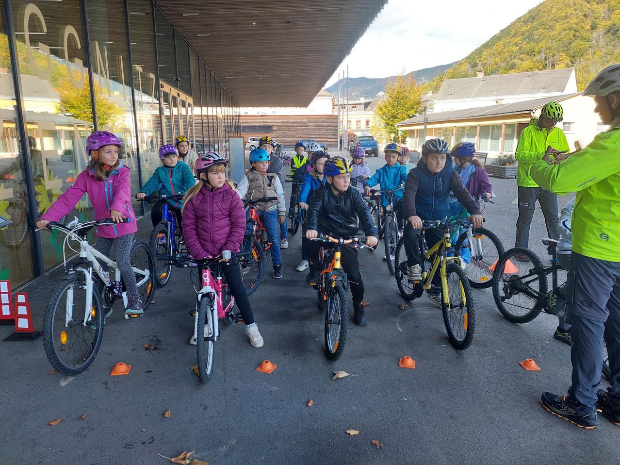 Eine Gruppe von Kindern ist auf Fahrrädern in einem Außenbereich versammelt und trägt Helme. Sie sind mit orangefarbenen Kegeln auf dem Boden aufgereiht. Dahinter steht ein Gebäude mit Glaswänden.