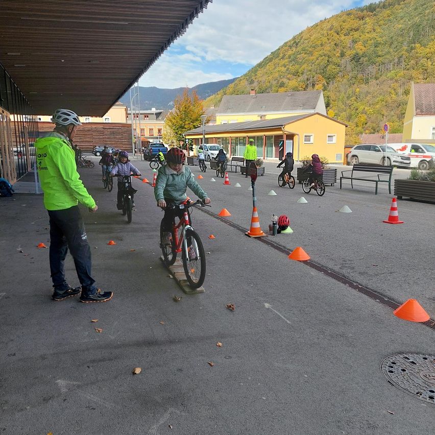 Eine Gruppe von Menschen übt das Radfahren auf einer asphaltierten Straße mit Verkehrskegel. Ein Mann führt einen Radfahrer in Grün. Dahinter stehen mehrere Gebäude.