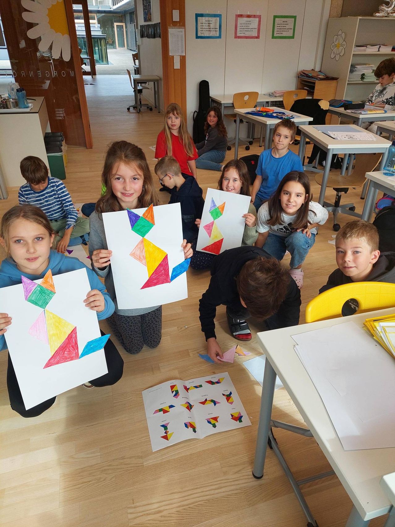 In einem Klassenzimmer sitzen mehrere Kinder auf dem Boden und halten bunte Papierausschnitte hoch. Sie sind in einer Bastelaktivität engagiert. Einige Kinder lächeln, andere schauen nach unten.
