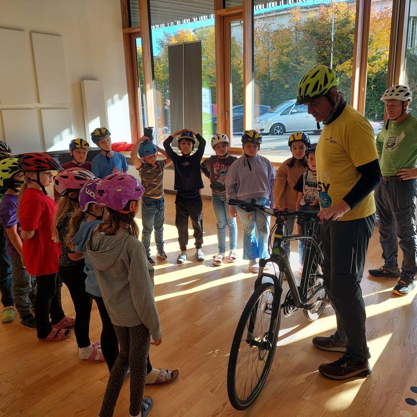 Eine Gruppe von Kindern mit Helmen steht um einen Mann herum, der etwas über ein Fahrrad erklärt. Sie befinden sich in einem Raum mit Holzböden und Glasfenstern.