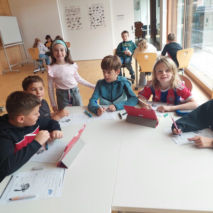 Ein Klassenzimmer mit Kindern, die an Tischen sitzen und Schreib- und Zeichenaktivitäten durchführen. Ein Mädchen in einem rosa Shirt steht zwischen ihnen, und mehrere Kinder haben Tablets auf ihren Schreibtischen.