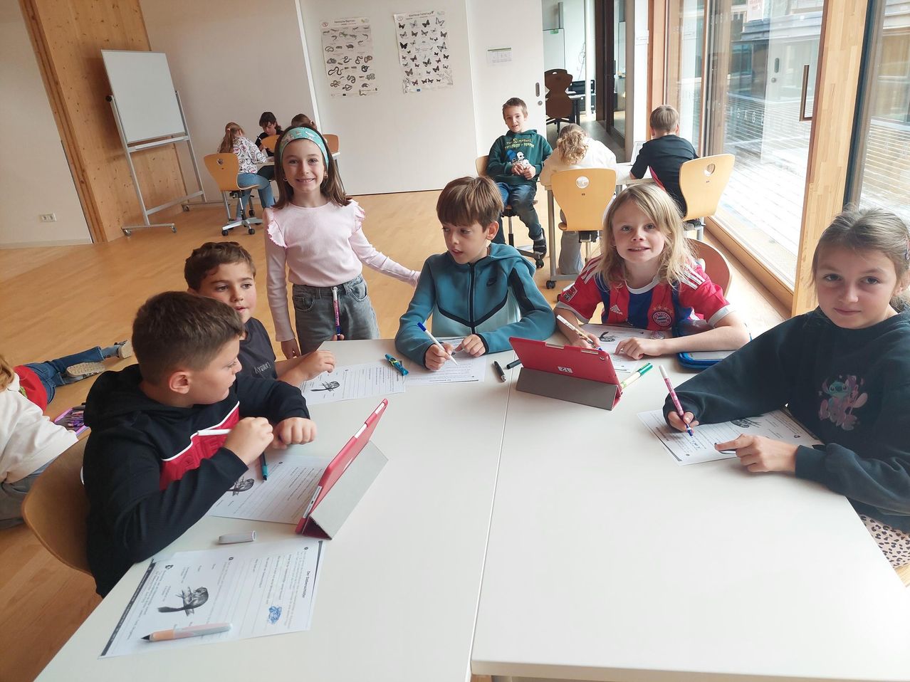 Ein Klassenzimmer mit Kindern, die an Tischen sitzen und Schreib- und Zeichenaktivitäten durchführen. Ein Mädchen in einem rosa Shirt steht zwischen ihnen, und mehrere Kinder haben Tablets auf ihren Schreibtischen.