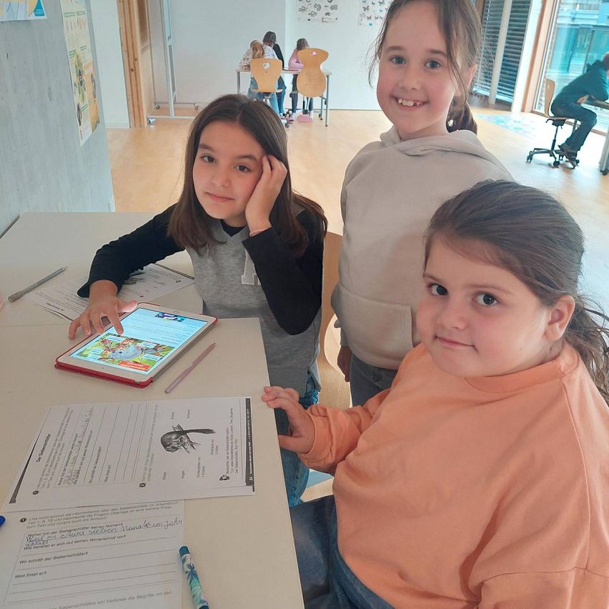 Drei junge Mädchen sitzen um einen Tisch mit einem Tablet, Bleistiften und Papieren. Sie befinden sich in einem Klassenzimmer mit einer weißen Wand und einem Holzboden. Hinter ihnen stehen Tische und Stühle.