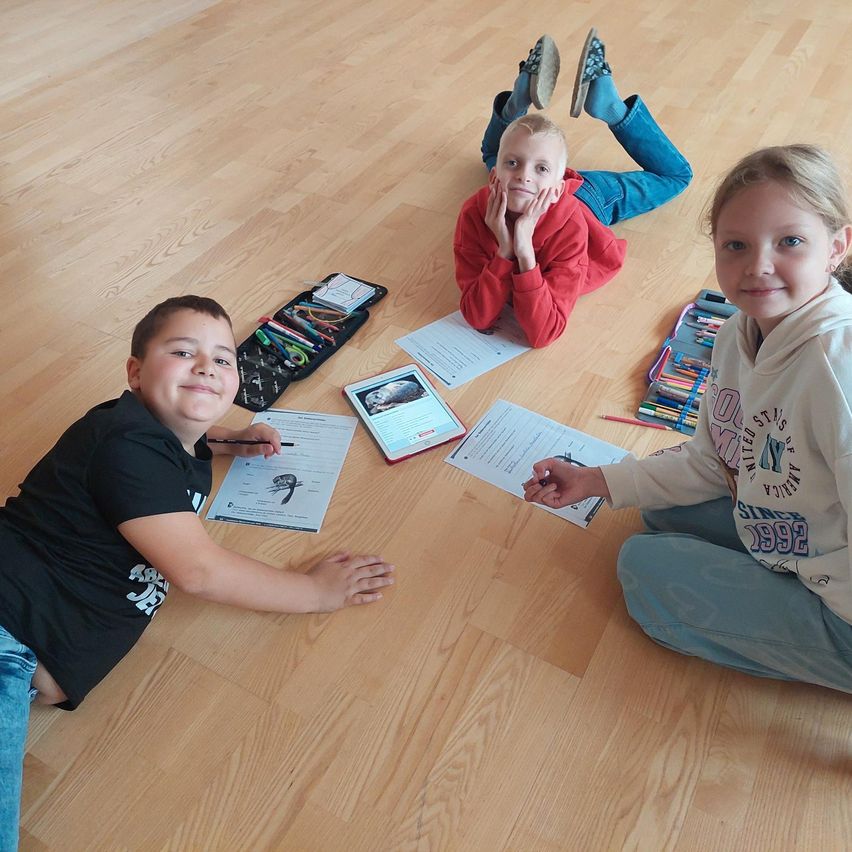 Drei Kinder sitzen auf einem Holzboden, wahrscheinlich in einem Klassenzimmer. Sie haben Papiere und Bleistifte vor sich. Der Junge links lächelt und hält einen Bleistift. Der Junge in der Mitte liegt auf dem Bauch mit den Händen im Gesicht. Das Mädchen rechts lächelt und hält einen Bleistift.