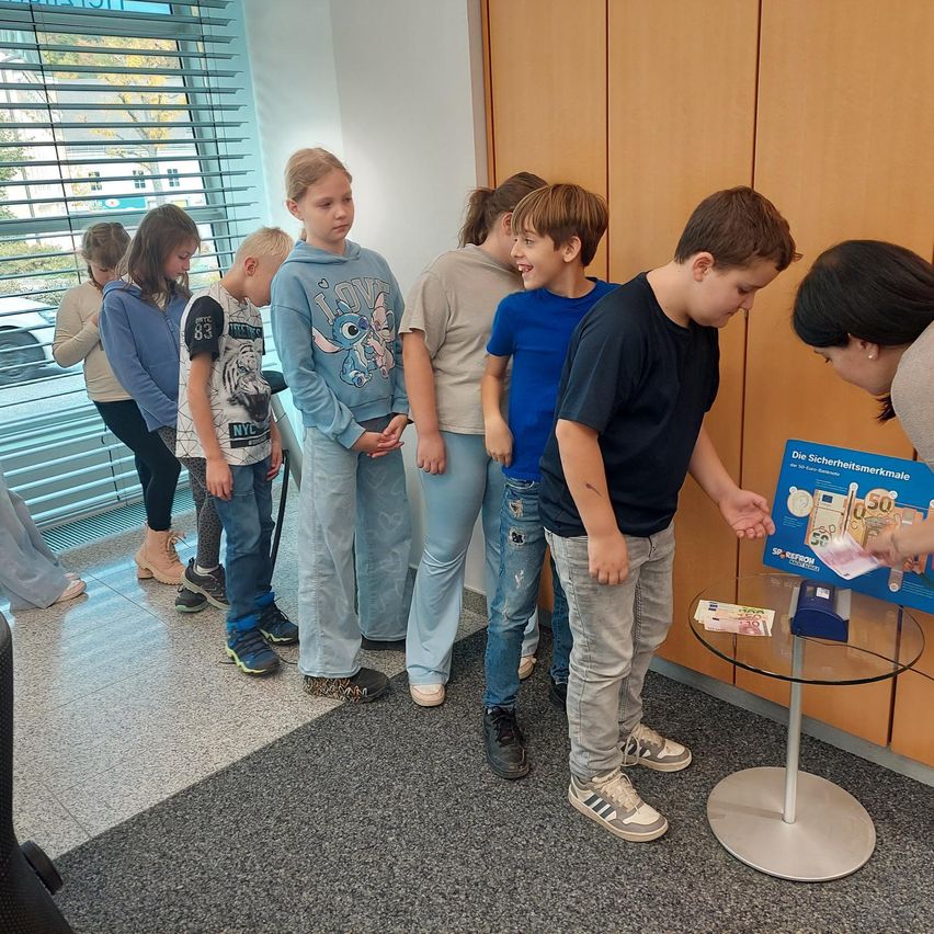 Eine Gruppe von Kindern steht um einen Tisch mit einem Geldautomaten, während eine Frau auf ein Poster zeigt. Sie lernen wahrscheinlich den Umgang mit Geld.