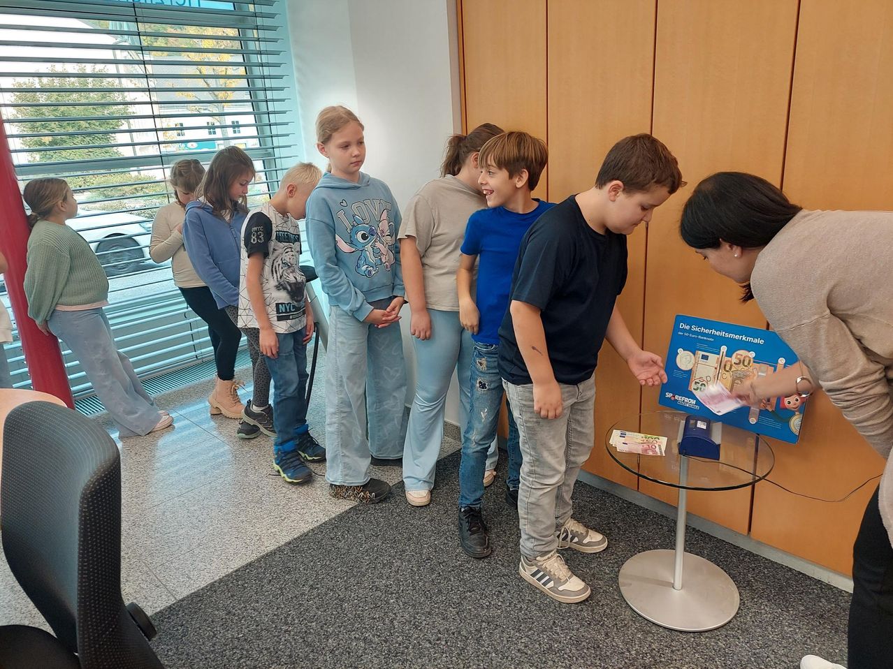 Eine Gruppe von Kindern steht um einen Tisch mit einem Geldautomaten, während eine Frau auf ein Poster zeigt. Sie lernen wahrscheinlich den Umgang mit Geld.