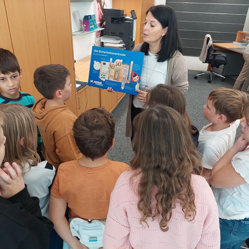 Eine Frau steht vor einer Gruppe von Kindern und hält ein blaues Plakat mit Bildern von Geld. Die Kinder schauen auf das Plakat und scheinen interessiert zu sein. Die Frau unterrichtet sie wahrscheinlich über Geld.