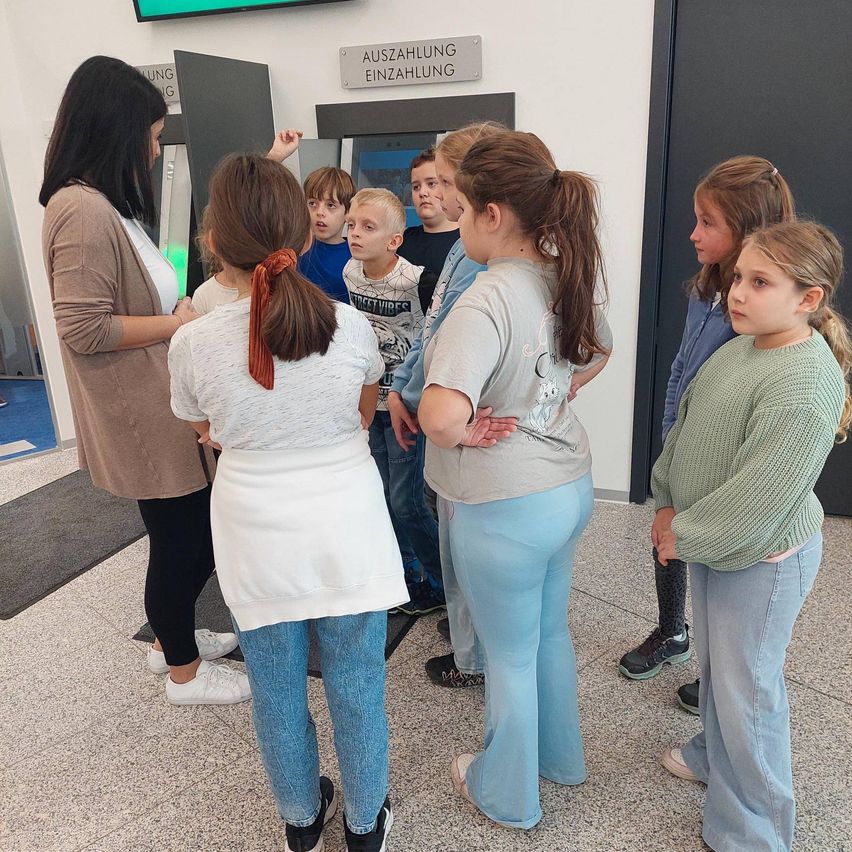 Eine Frau führt eine Gruppe von Kindern in einem Flur. Sie stehen um eine Tür herum, die mit 'Auszahlung Einzahlung' beschriftet ist. Die Kinder tragen Freizeitkleidung. Der Boden ist glänzend, und ein Teppich befindet sich in der Nähe der Tür.