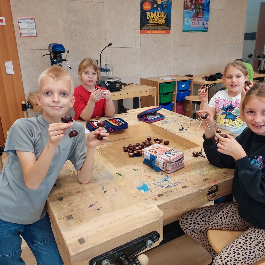 Vier Kinder sitzen um einen Holztisch in einem Klassenzimmer. Sie halten eine Kastanie und einen Stock, und der Tisch ist mit Kastanien gefüllt. Es gibt Poster an der Wand und eine Bohrmaschine auf der linken Seite.