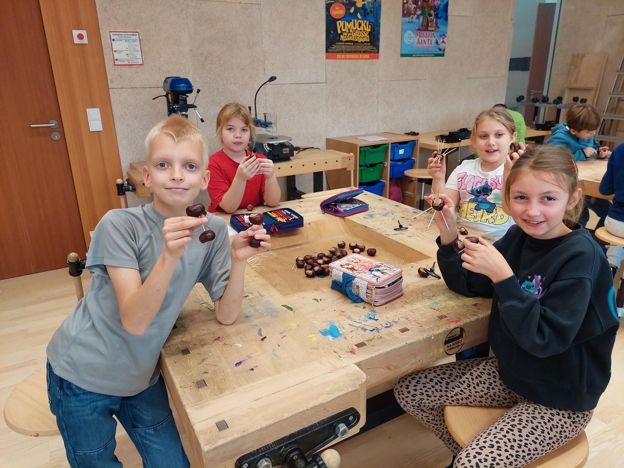 Vier Kinder sitzen um einen Holztisch in einem Klassenzimmer. Sie halten eine Kastanie und einen Stock, und der Tisch ist mit Kastanien gefüllt. Es gibt Poster an der Wand und eine Bohrmaschine auf der linken Seite.