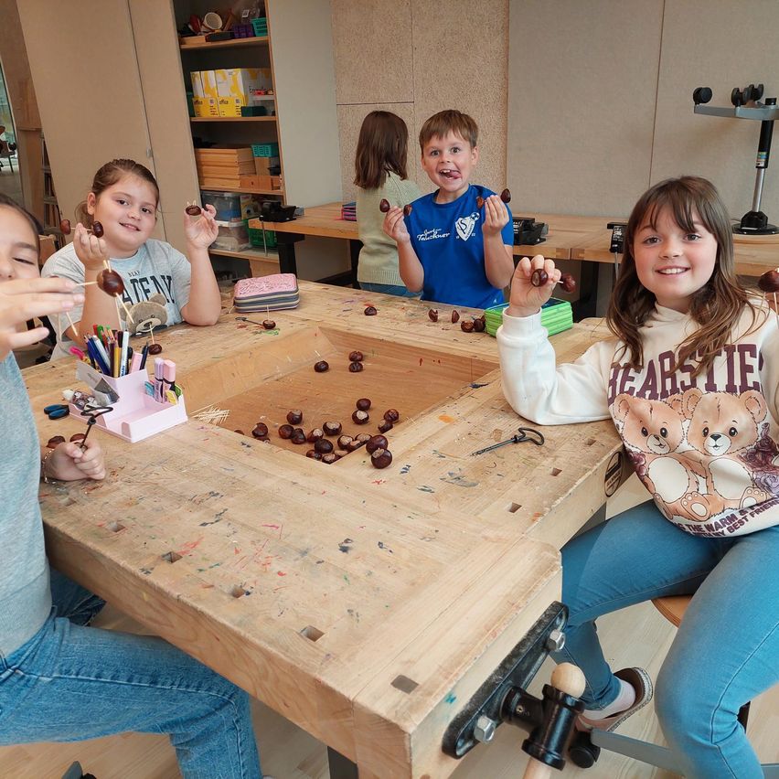 Eine Gruppe von Kindern sitzt um einen Holztisch herum, jeder hält Kastanien auf Stöcken und lächelt, während sie an einer Bastelaktivität teilnehmen.