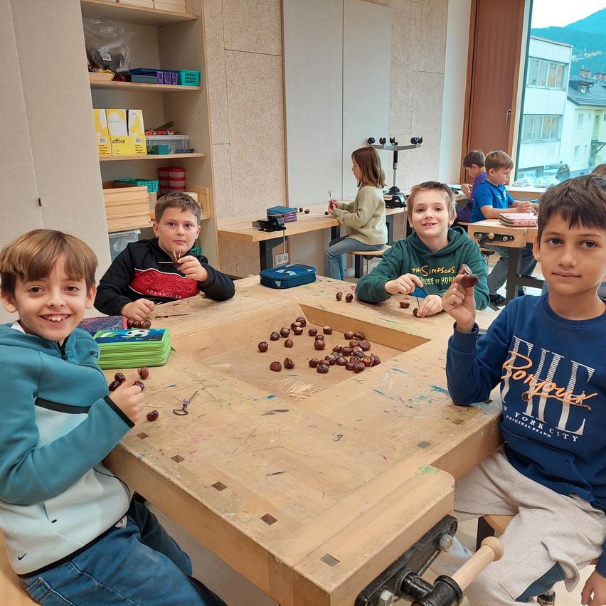 Mehrere Kinder sitzen an einem Holztisch und beschäftigen sich mit einer Bastelaktivität. Sie halten Gegenstände in den Händen und lächeln. Hinter ihnen sitzt eine Frau an einem anderen Tisch und arbeitet ebenfalls an einem Bastelprojekt. Der Raum hat Regale mit verschiedenen Gegenständen und ein Fenster mit Blick auf Gebäude.