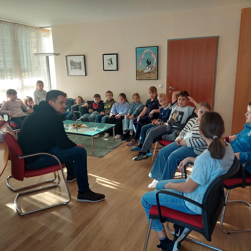 Eine Gruppe von Kindern und einem Mann sitzen auf Stühlen in einem Raum mit Holzböden. Der Mann sitzt vorne, und die Kinder sitzen um einen Tisch herum.