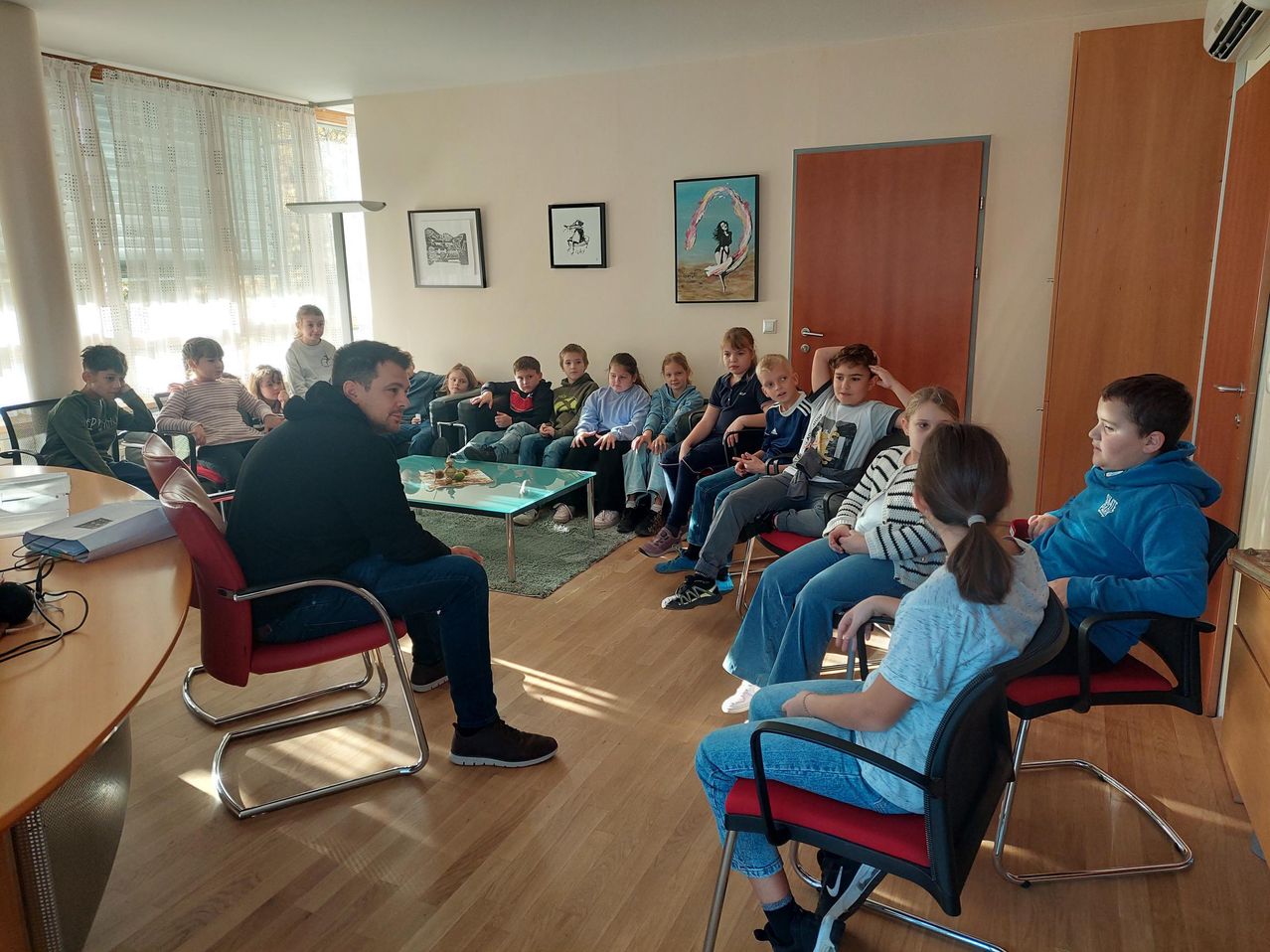 Eine Gruppe von Kindern und einem Mann sitzen auf Stühlen in einem Raum mit Holzböden. Der Mann sitzt vorne, und die Kinder sitzen um einen Tisch herum.