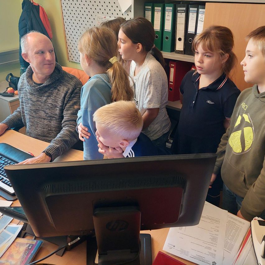 Eine Gruppe von Kindern und einem erwachsenen Mann versammelt sich um einen Computer. Der Mann sitzt und die Kinder stehen und schauen auf den Bildschirm.