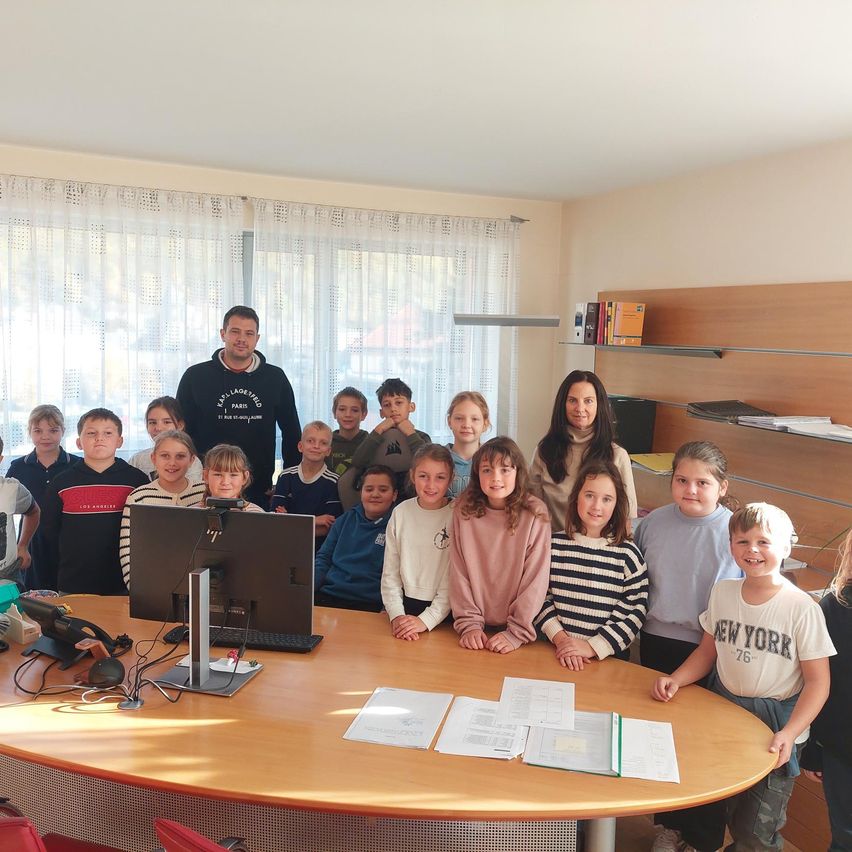 Eine Gruppe von Kindern und zwei Erwachsenen versammelt sich in einem Raum. Die Kinder sitzen um einen Tisch mit Papieren und einem Computer. Dahinter stehen eine Frau und ein Mann in der Nähe eines Fensters mit Vorhängen.