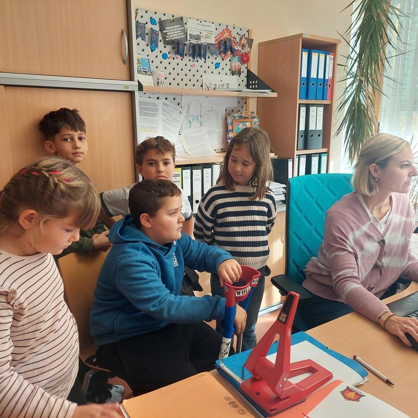Eine Gruppe von Kindern und eine Frau befinden sich in einem Büro. Die Kinder sind um eine rote Maschine herum versammelt. Die Frau sitzt auf einem Stuhl an einem Schreibtisch.