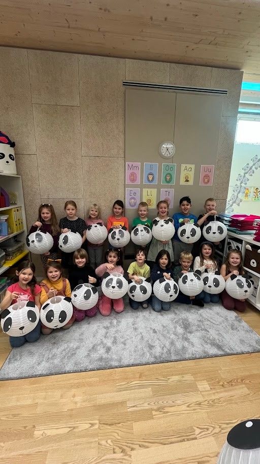 Eine Gruppe von Kindern in einem Klassenzimmer posiert mit Panda-Lampions. Der Raum hat einen Teppich, Regale und Alphabet-Poster an der Wand.