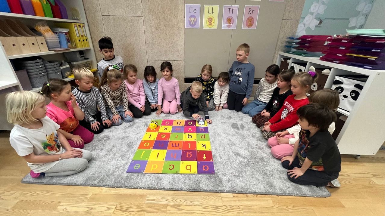 Eine Gruppe von Kindern sitzt im Kreis um einen bunten Alphabet-Teppich auf dem Boden, wobei einige Buchstaben hervorgehoben sind. Dahinter befinden sich Wände mit Alphabetbuchstaben und Bildern von Tieren.