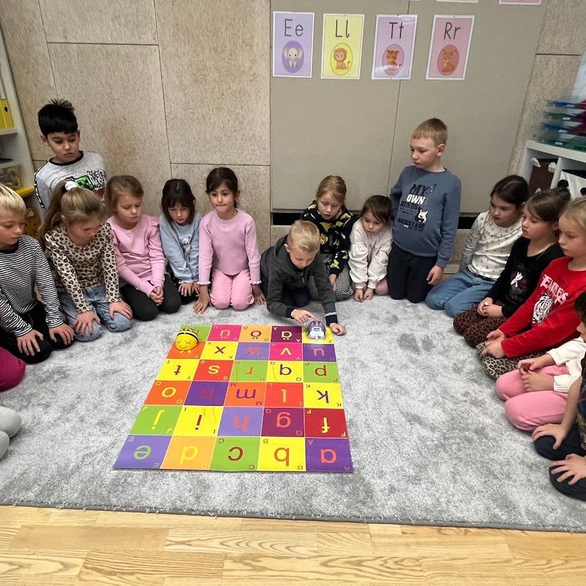 Eine Gruppe von Kindern sitzt im Kreis um einen bunten Alphabet-Teppich auf dem Boden, wobei einige Buchstaben hervorgehoben sind. Dahinter befinden sich Wände mit Alphabetbuchstaben und Bildern von Tieren.