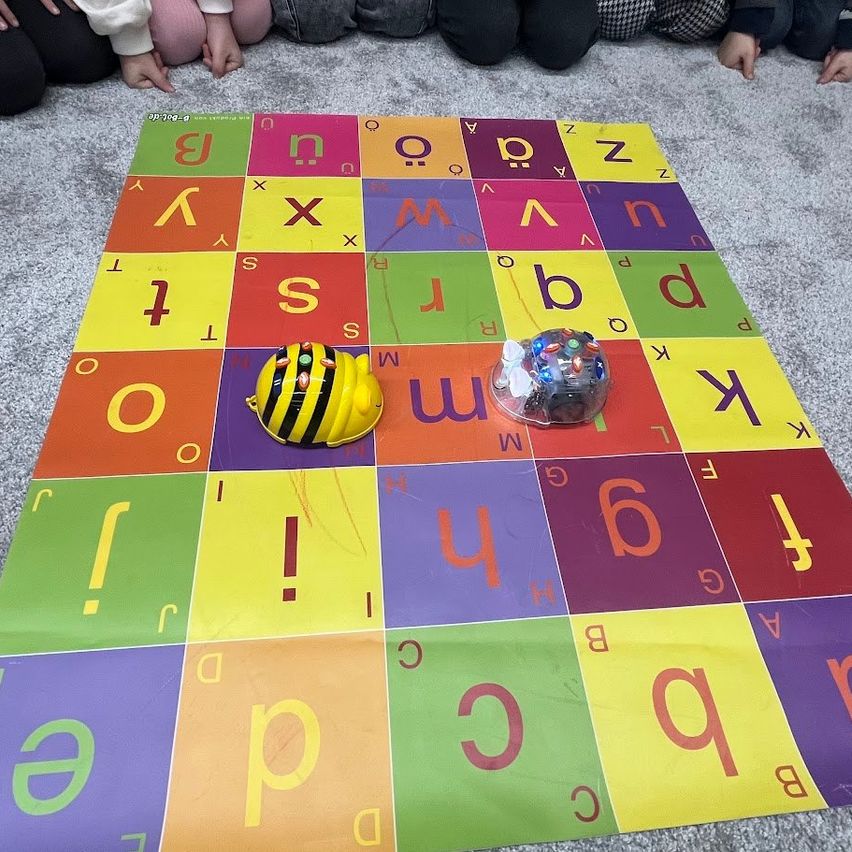 Ein bunter Lernteppich mit Buchstaben und Zahlen liegt auf dem Boden. Zwei Spielzeuge sind darauf platziert, eines sieht aus wie eine Biene und das andere wie ein Roboter.