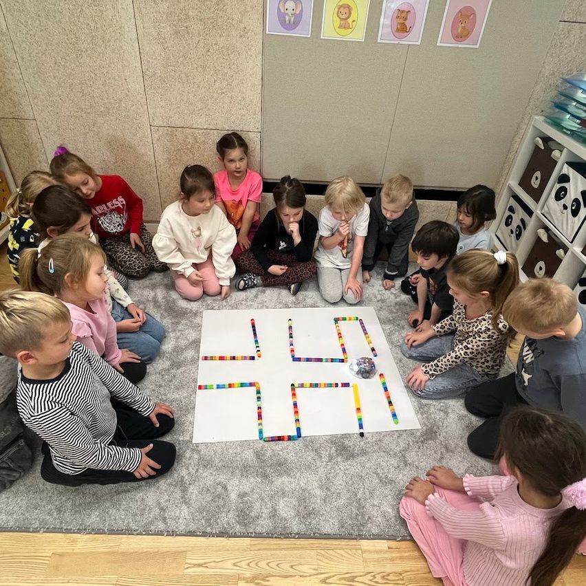 Eine Gruppe von Kindern sitzt auf dem Boden in einem Klassenzimmer. Sie sind um ein weißes Papier mit farbenfrohen Perlen in einem Muster herum versammelt. Die Kinder tragen Freizeitkleidung und scheinen etwas auf dem Papier zu beobachten. Der Raum hat eine weiße Wand mit Tierbildern und einem Holzboden.