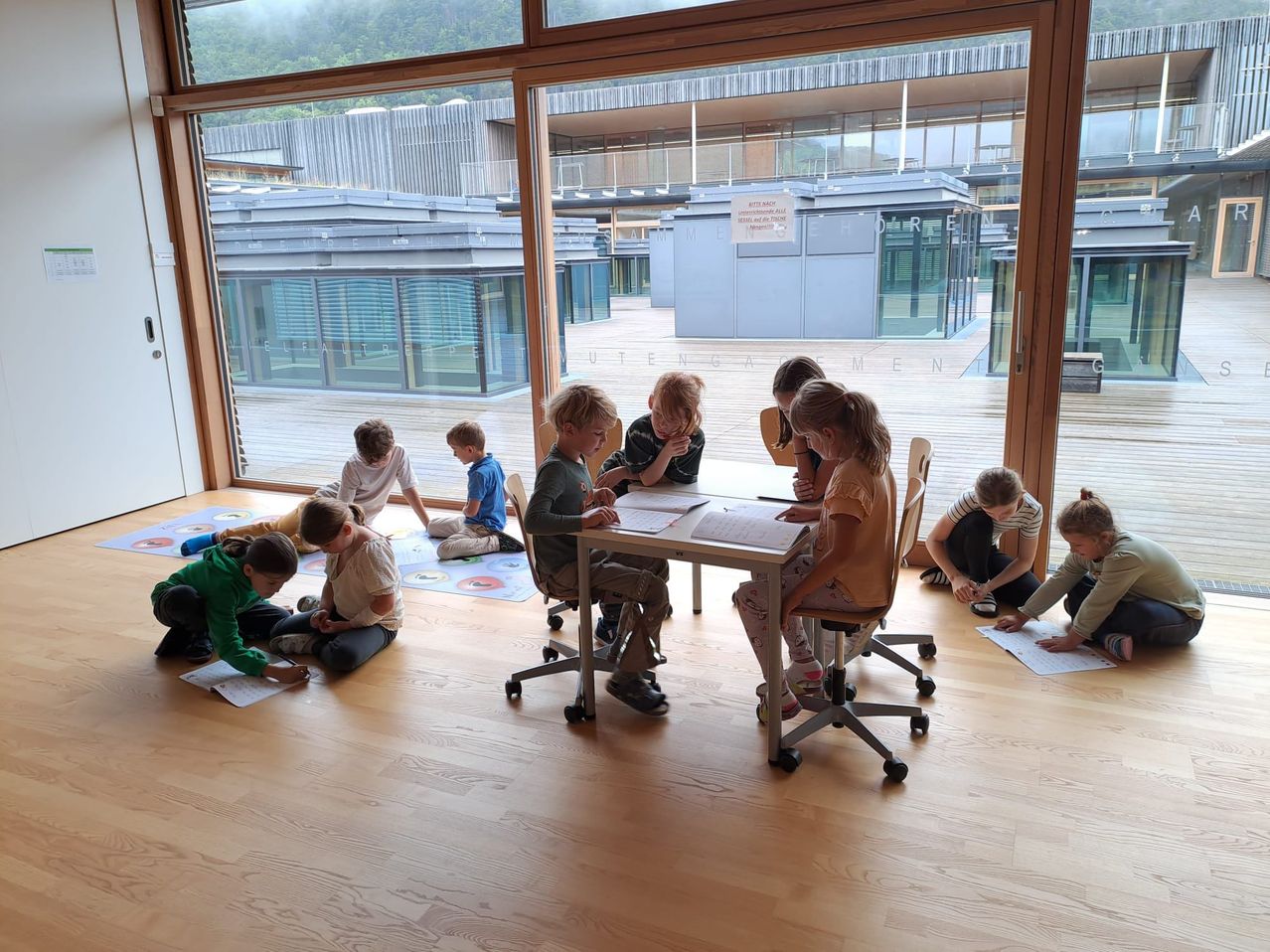 Ein Klassenzimmer mit Kindern, die auf dem Boden und Stühlen sitzen und einer Aktivität nachgehen. Ein Tisch hält Papiere und das Zimmer verfügt über große Fenster mit Blick nach draußen.