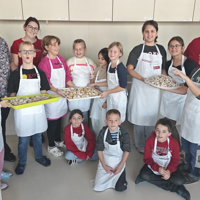 Eine Gruppe von Kindern und Erwachsenen in einer Küche, alle mit Schürzen bekleidet, halten Tabletts mit Keksen. Einige stehen, andere sitzen. Die Frau in der Mitte hält ein Tablett mit Keksen.