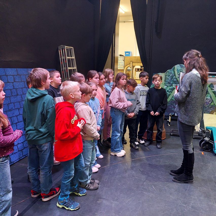 Eine Gruppe von Kindern und einer Frau stehen auf der Bühne und proben möglicherweise für ein Theaterstück. Die Kinder hören der Frau zu, die ein Buch in der Hand hält.
