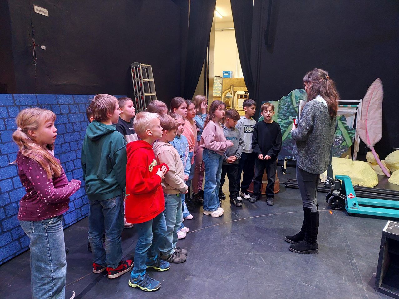 Eine Gruppe von Kindern und einer Frau stehen auf der Bühne und proben möglicherweise für ein Theaterstück. Die Kinder hören der Frau zu, die ein Buch in der Hand hält.