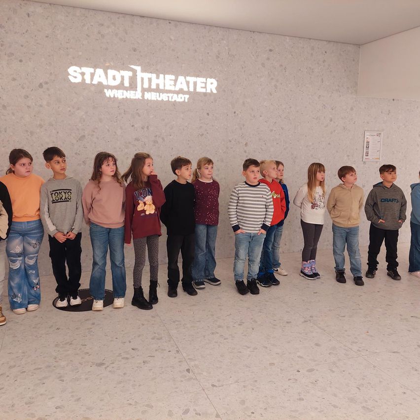 Eine Gruppe von Kindern steht in einer Reihe vor einer Wand mit einem Schild, das 'Stadt Theater Wiener Neustadt' steht. Sie alle schauen in die gleiche Richtung.