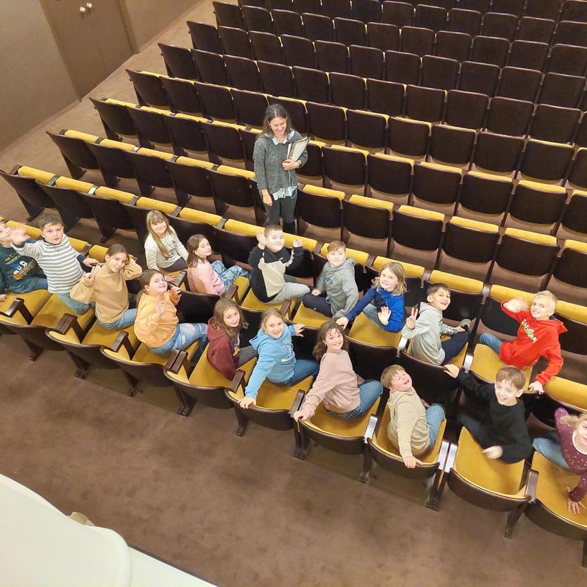 Eine Frau steht hinten in einem Auditorium, vor ihr sitzen Kinder in den Reihen der Stühle. Die Kinder lächeln und machen Gesten.