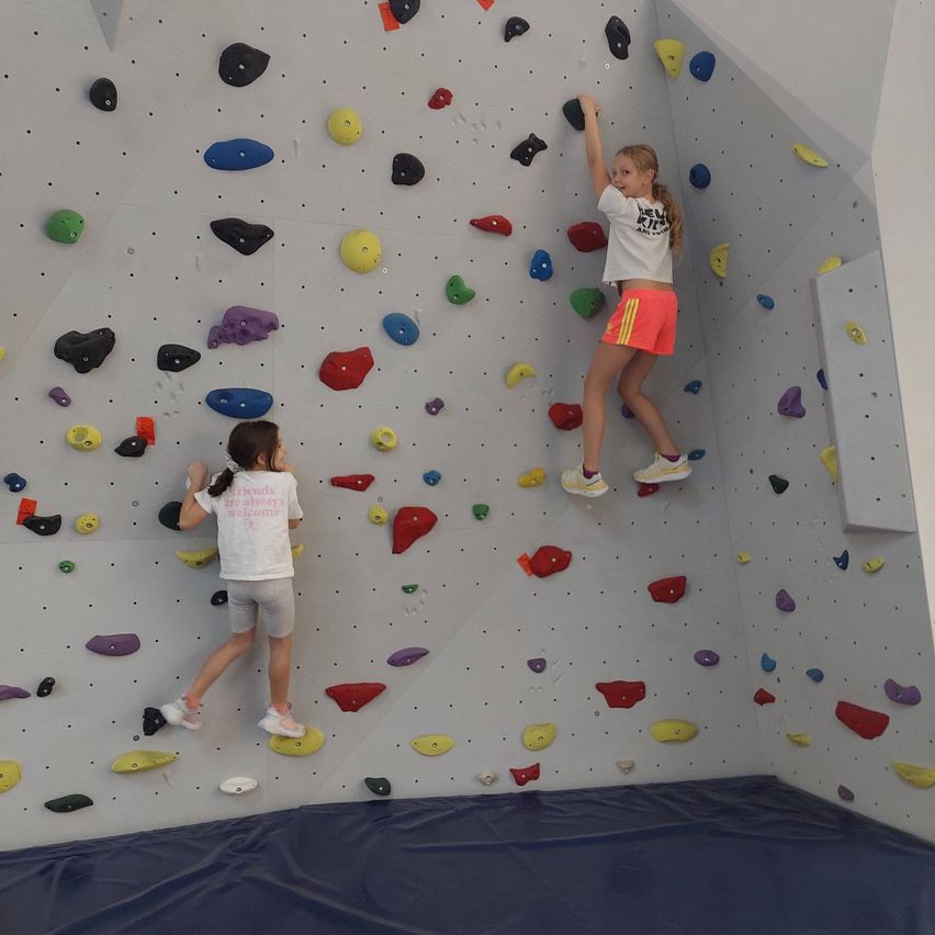 Zwei junge Mädchen klettern an einer Kletterwand mit farbigen Griffen. Die Wand ist mit einer weißen Beschichtung bedeckt.