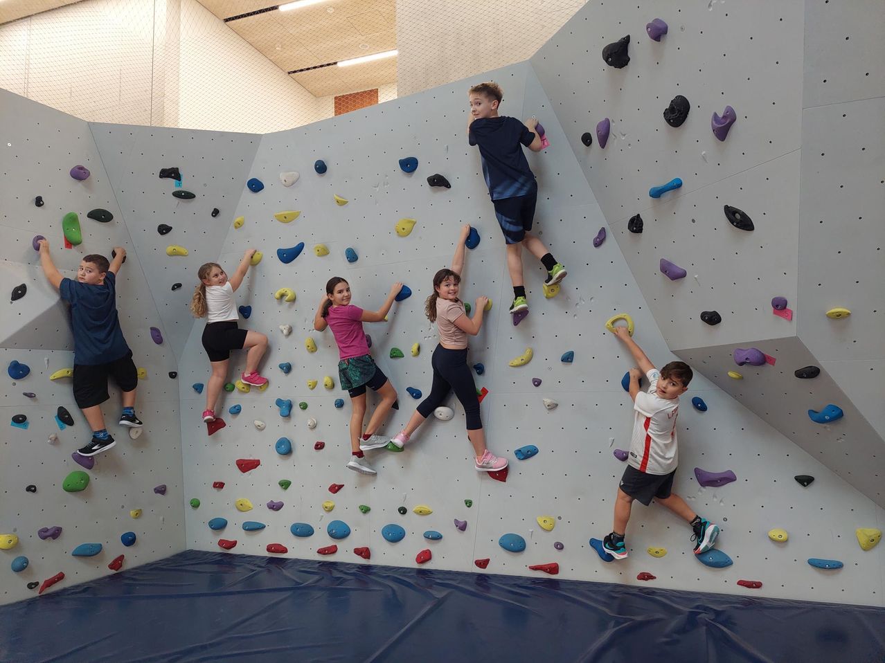 Eine Gruppe von Kindern klettert an einer bunten Kletterwand, einige mit den Händen und andere mit den Füßen. Sie tragen Turnschuhe und Shorts. Die Wand hat verschiedene farbige und geformte Griffe.