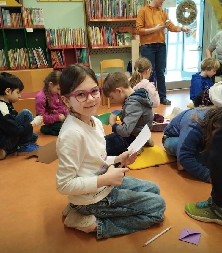 Kinder sitzen in einer Bibliothek auf dem Boden, einige lesen Bücher. Ein Mädchen vorne lächelt und hält ein Buch und einen Stift. Dahinter steht eine Frau, vielleicht eine Lehrerin.