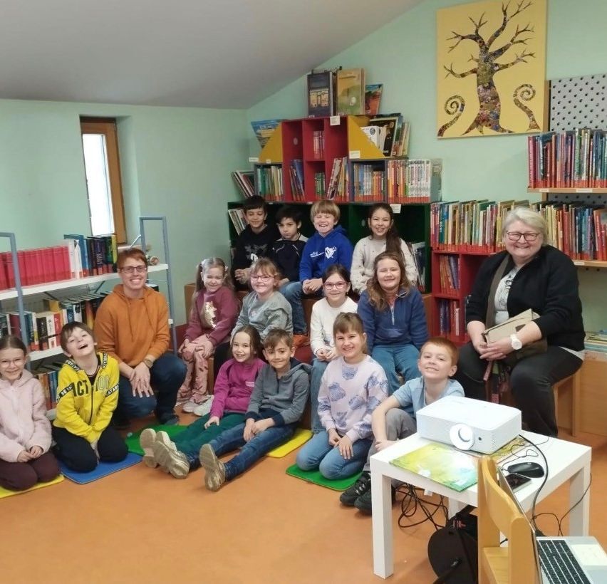 Eine Gruppe von Kindern und zwei Erwachsenen sitzen auf dem Boden in einer Bibliothek. Ein Projektor steht auf einem Tisch, und Regale mit Büchern sind im Hintergrund.