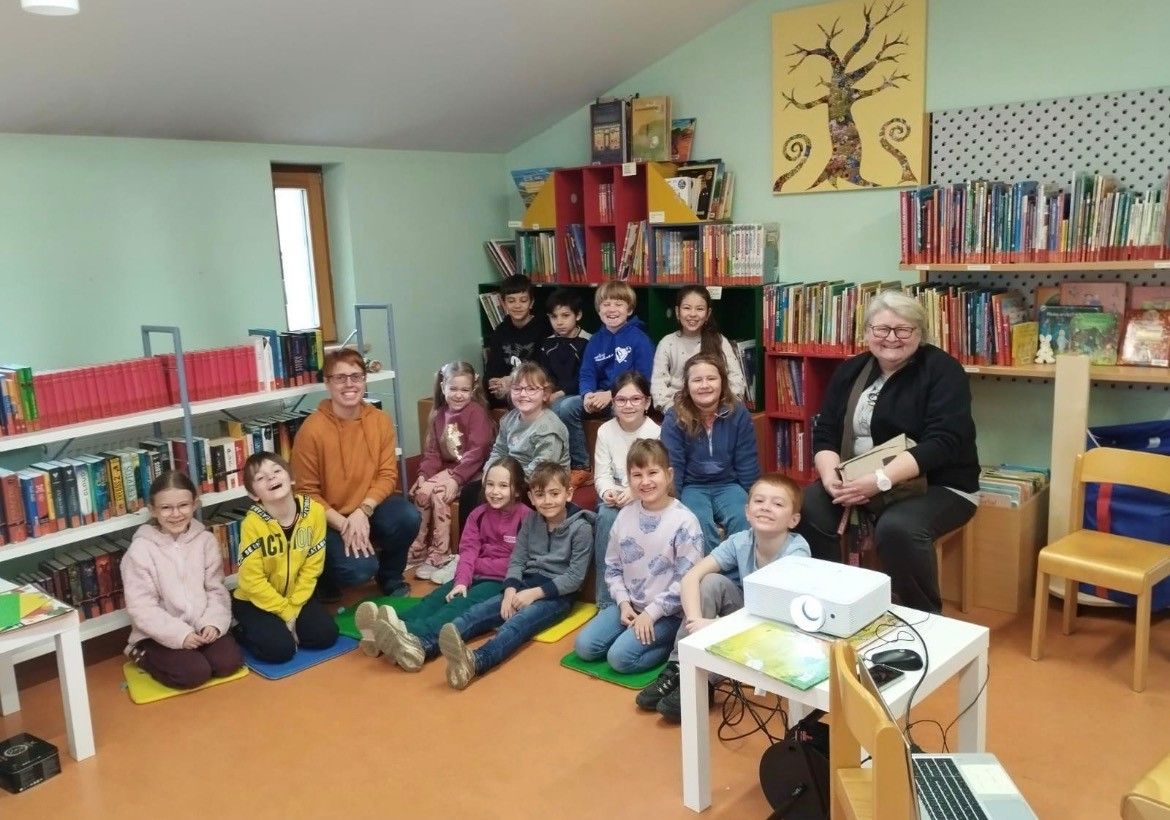 Eine Gruppe von Kindern und zwei Erwachsenen sitzen auf dem Boden in einer Bibliothek. Ein Projektor steht auf einem Tisch, und Regale mit Büchern sind im Hintergrund.