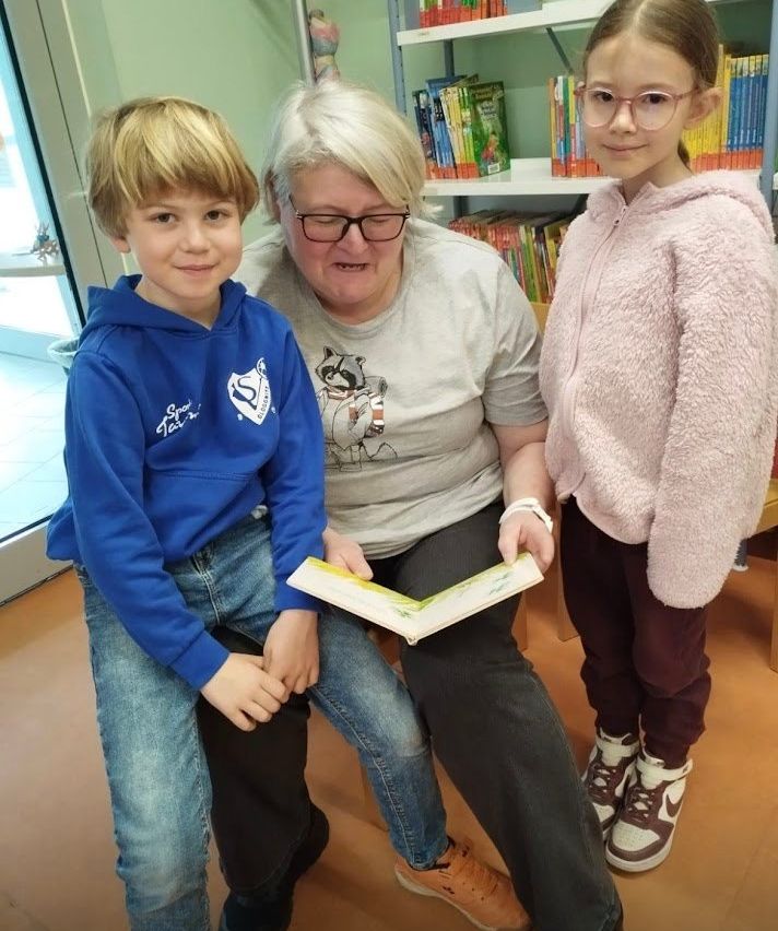 Eine ältere Frau liest zwei Kindern ein Buch vor, eines trägt eine blaue Kapuzenjacke und das andere eine rosa Jacke. Sie sind in einem Raum mit Bücherregalen und einer Glastür im Hintergrund.